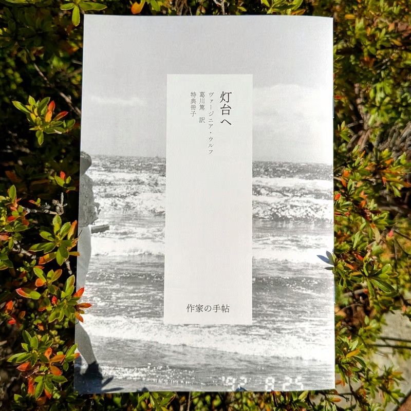 作家の手帖から刊行される、ヴァージニア・ウルフ『灯台へ』（葛川篤訳）予約特典冊子の書影画像。冊子の現物を植栽のなかで撮影した写真です。冊子の表紙には波打ち際の景色の写真が白黒で印刷されています。