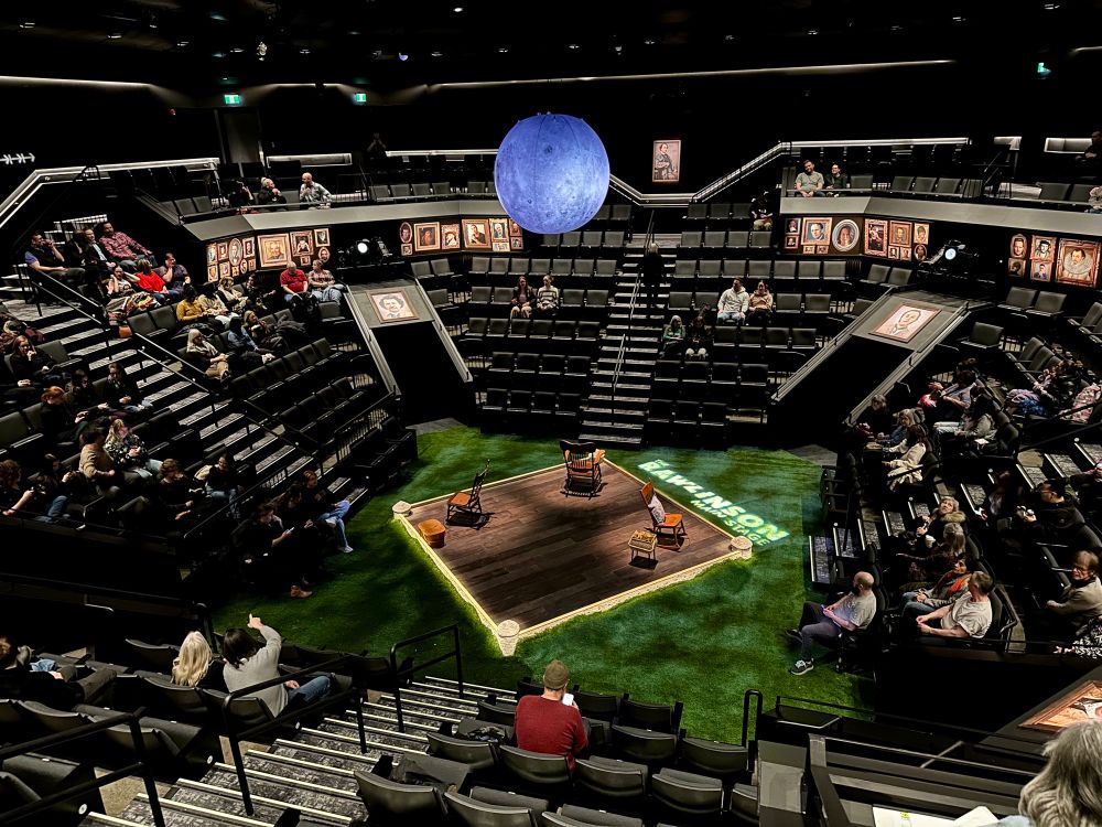 Panoramic view of a theatre in the round. The globe theatre in Regina stretches out across the entire frame, with seating on all sides with a smattering of audience. On the stage is set of green grass astroturf, small walls and turrets of a fort. Inside the “fort” is a wooden floor, three rocking chairs and three sewing baskets. The rocking chairs are lit with a spot light each and the walls the fort are glowing. 
