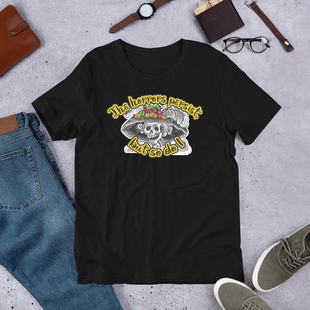 Black t-shirt with a skeleton wearing a large hat with colourful flowers, surrounded by the text "The horrors persist, but so do I". 