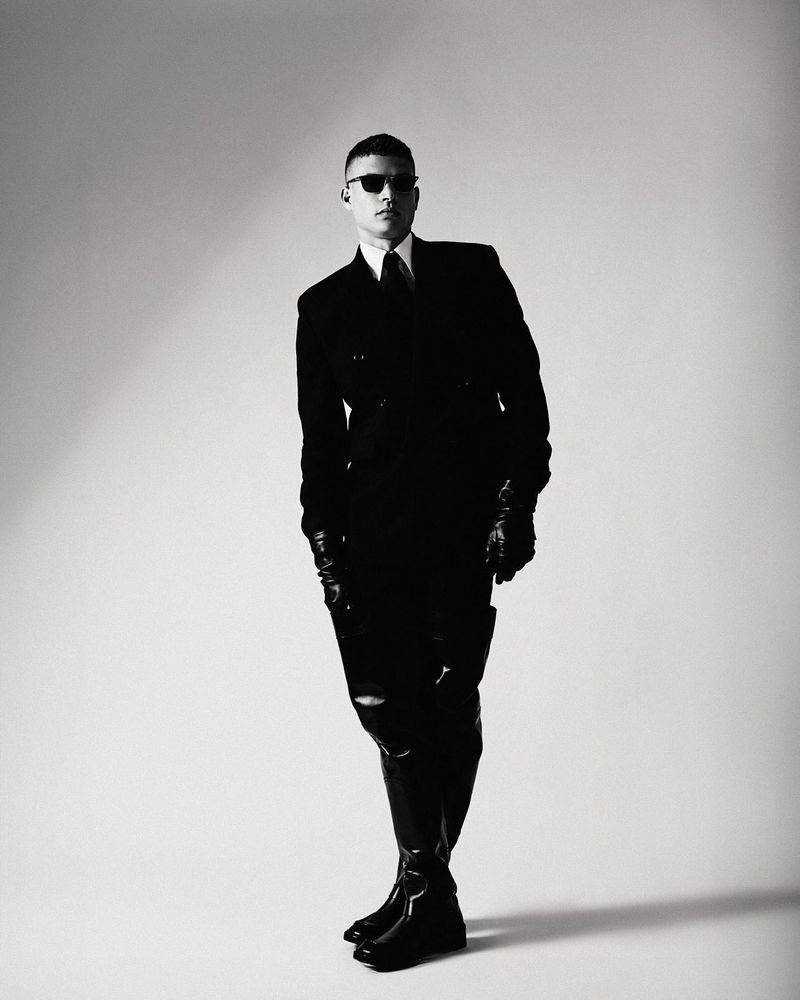 A man dressed in a sharp black suit with leather gloves and sunglasses, standing with a confident pose in a minimalist studio setting, photographed in black and white.
