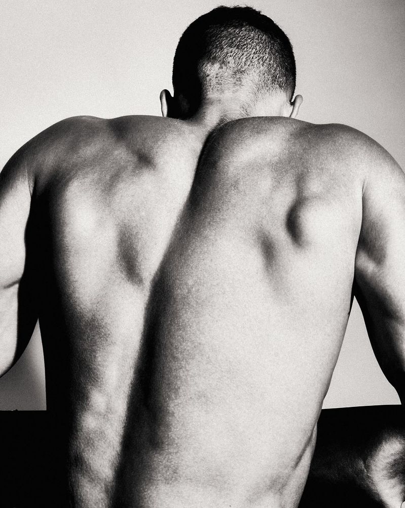 A black-and-white photo capturing the muscular, bare back of a man, showing his defined shoulders and short haircut, with dramatic lighting emphasizing his physique.

