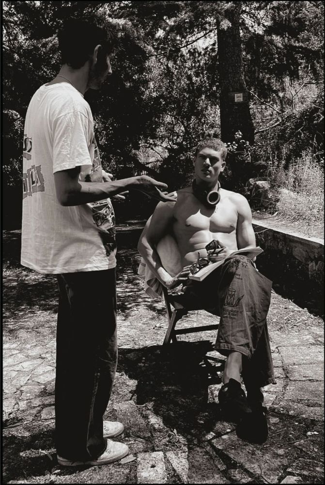 A black-and-white outdoor photo showing a shirtless man sitting on a folding chair, wearing loose cargo pants and headphones around his neck, holding sunglasses and a notebook. Another man stands in front of him mid-gesture, possibly in conversation. The setting is shaded by trees with a rustic, stone-paved ground.
