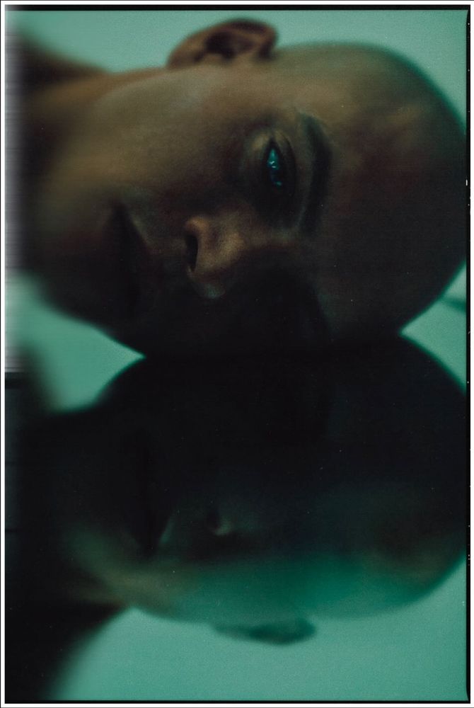 A dramatic, low-light close-up photo of a man's face resting sideways on a reflective surface, possibly glass. The dim, teal-tinted lighting creates a moody, introspective atmosphere, and his face is partially duplicated in the reflection.