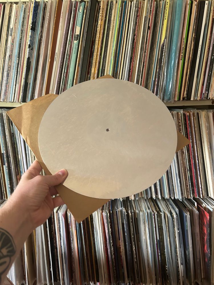Me holding up a blank aluminum disc from the Virginia Folklore Society recordings against the backdrop of some of the LPs in our living room. The blank disc is shiny and reflects the ceiling of the room as a white/silver circle with a hole in the middle.