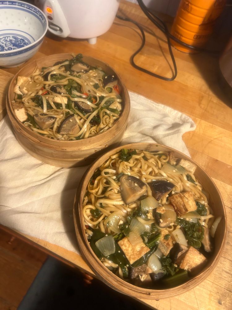 Two steamer baskets loaded with mustard greens onions tofu mushrooms and lightly seasoned noodles.