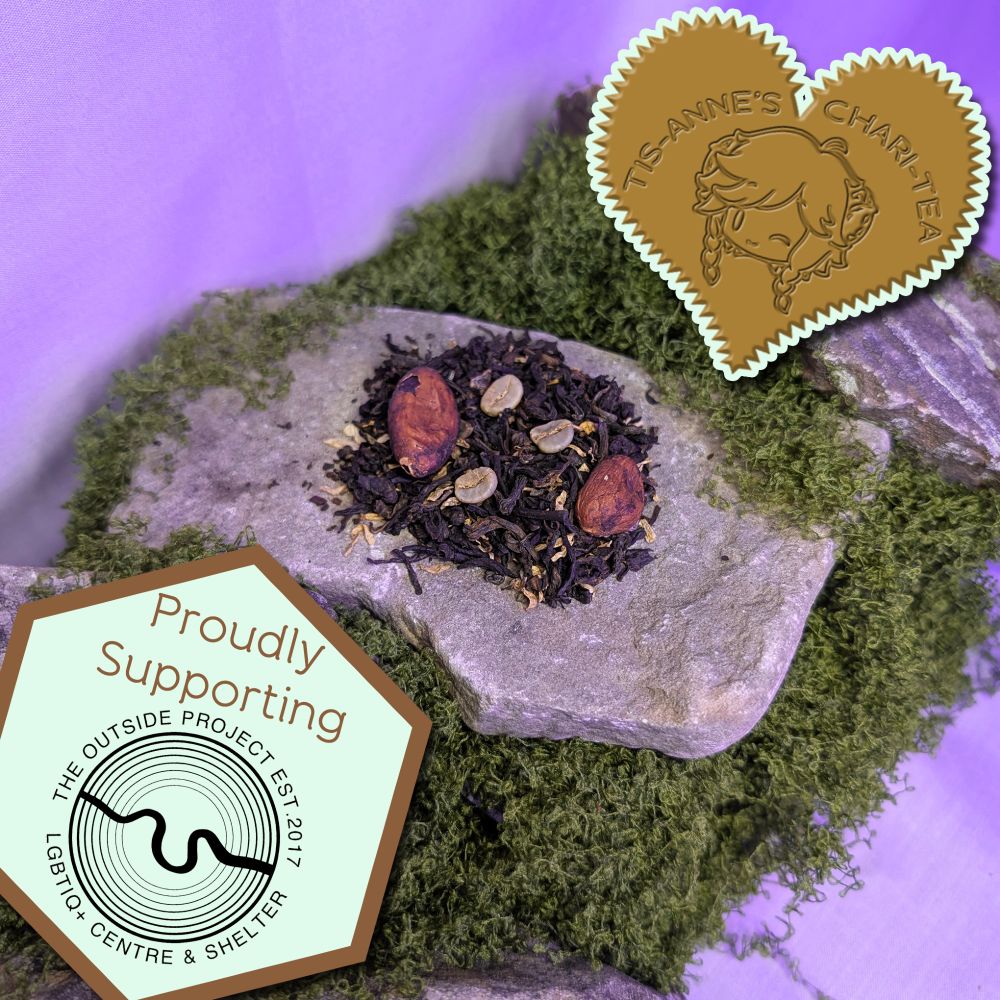 A small mound of tea leaves, with coffee and cocoa beans blended into it on top of a piece of grey slate. The slate itself is perched onto a heap of moss, and is surrounded by other pieces of slate. A heart-shaped badge in one corner reads "Tis-Anne's Chari-Tea", and another emblem reads "Proudly Supporting" with the logo for The Outside Project.