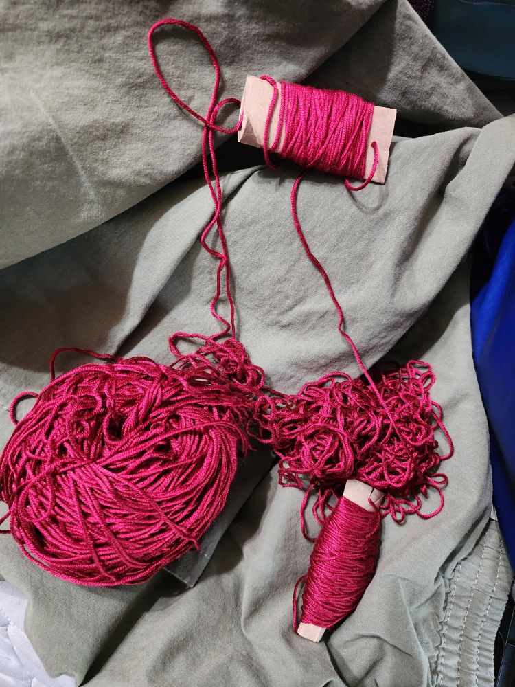 Tangled ball of shiny red yarn with two ends wrapped around cardboard scrap. It is in the process of being untangled. Sitting on top of some olive green shorts. 