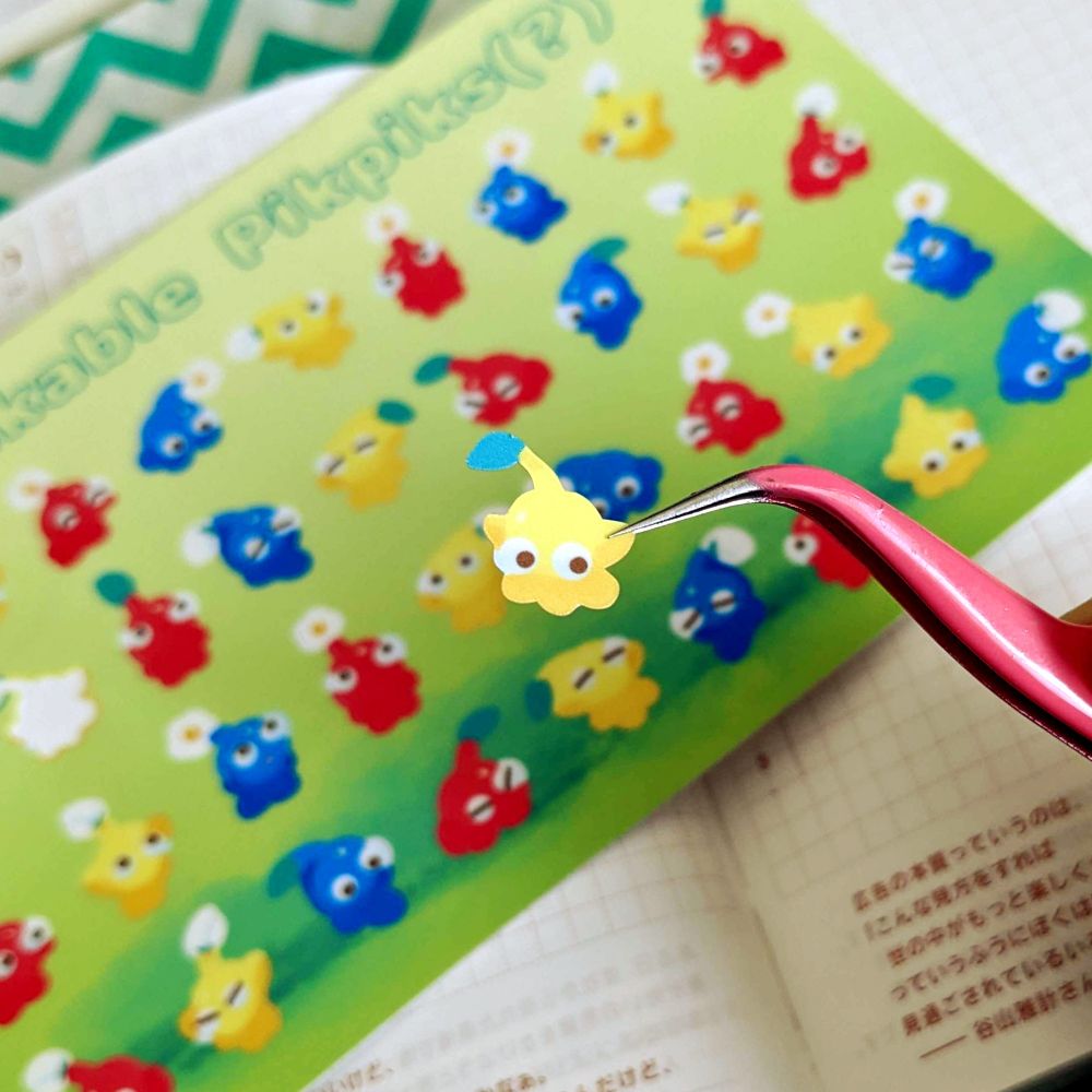 A tweezer holding up a yellow Pikmin sticker about half an inchtall. The sticker sheet of 35 other Pikmins are seen in the background on top of a journal.