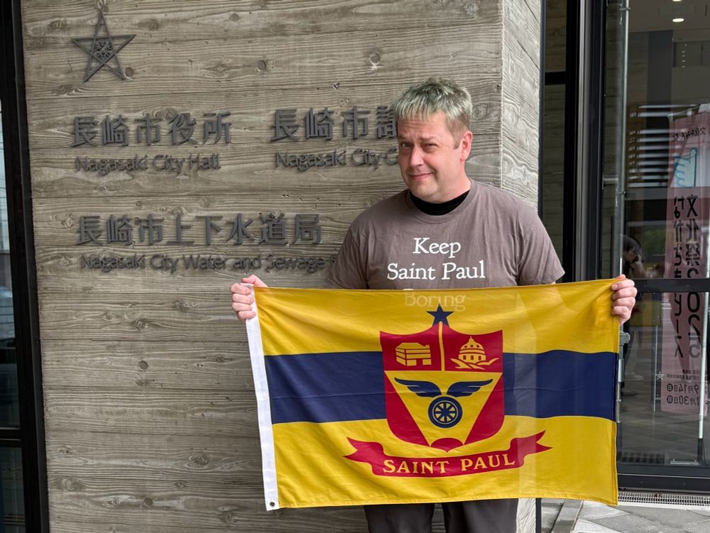 Me in front of Nagasaki City Hall, holding the Saint Paul city flag and wearing my “Keep Saint Paul Boring” shirt