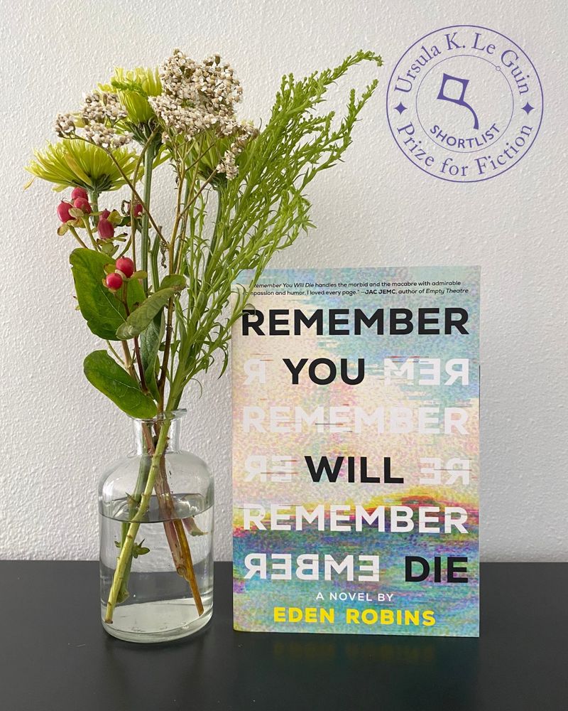 A photograph of Eden Robins' novel Remember You Will Die, which rests on a black metal shelf next to a small glass vase full of greenery: red berries, green and white flowers, and a shaggy sprig of foliage.