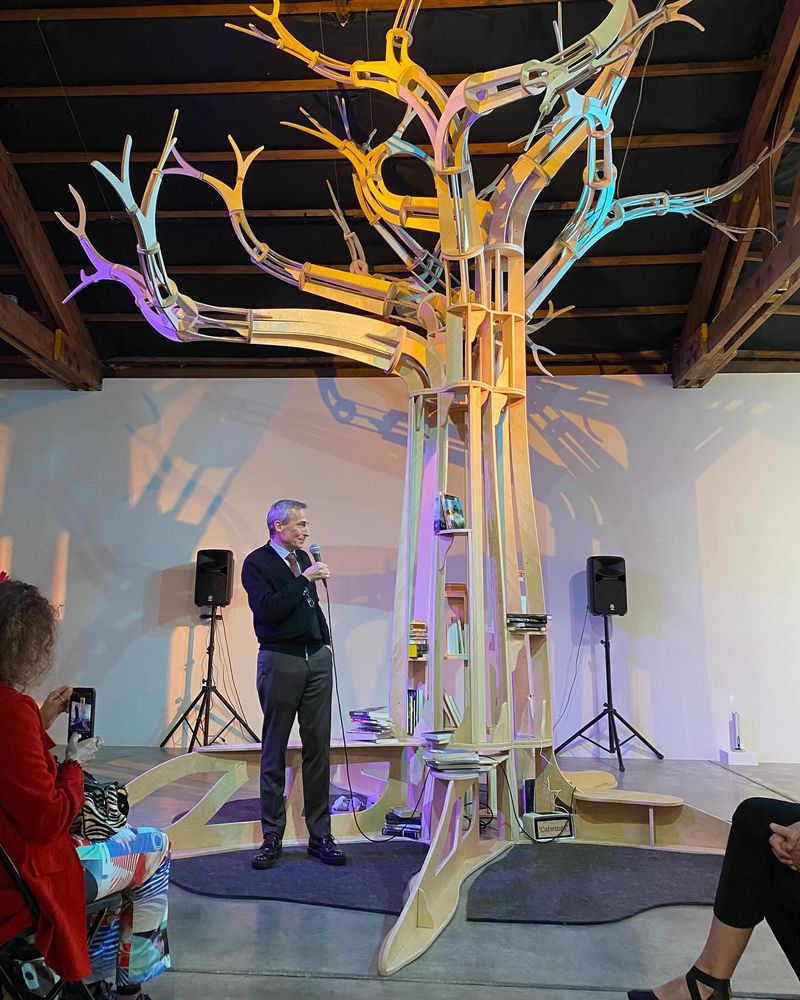 Theo Downes-Le Guin stands under the replica oak tree at the center of the A Larger Reality exhibition, introducing the exhibition on its opening night. The tree trunk has small bookshelves which hold copies of Ursula's books for visitors to read.