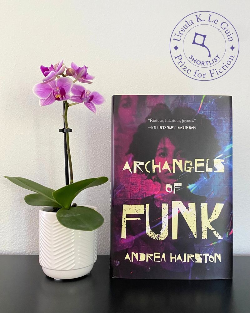 A copy of Archangels of Funk by Andrea Hairston, photographed next to a small purple orchid, sitting on a dark flat surface against a white background.