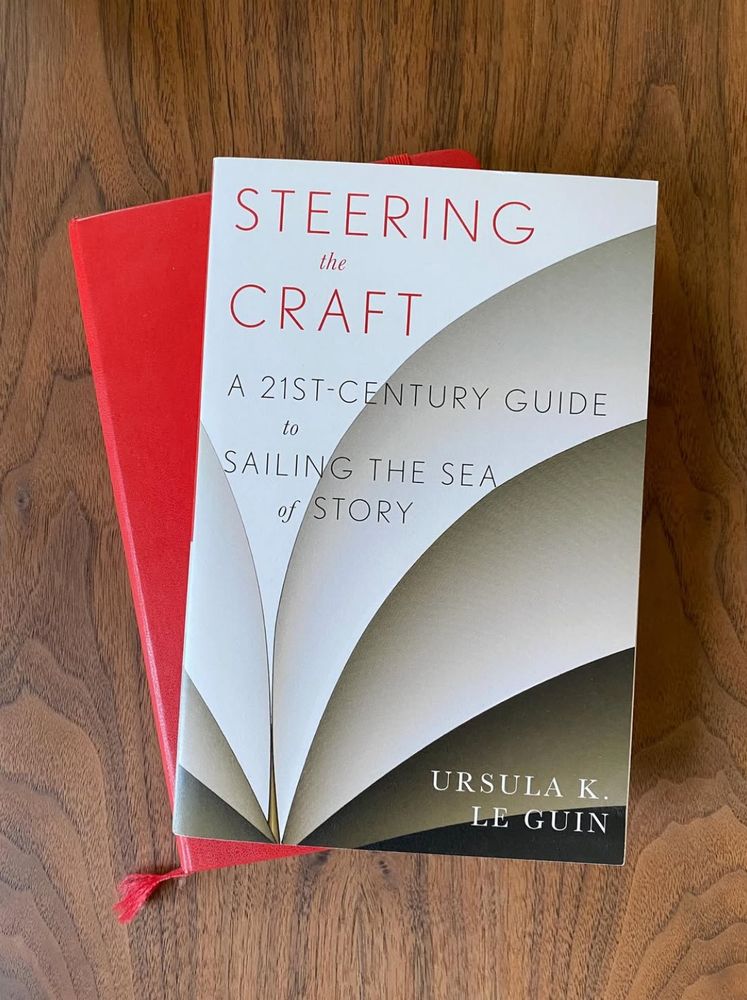 A copy of Steering the Craft sits on top of a red Moleskine notebook; both are on a wooden table