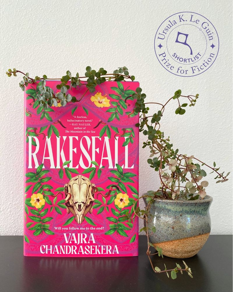 A copy of Rakesfall—the hot pink cover of which depicts a skull surrounded by flowers and vines—sits on a black surface against a white wall. A small pot next to it contains a vine-like plant, which is draped across the top of the book. The Ursula K. Le Guin Prize for Fiction Shortlist medal is in the upper right corner of the photo.