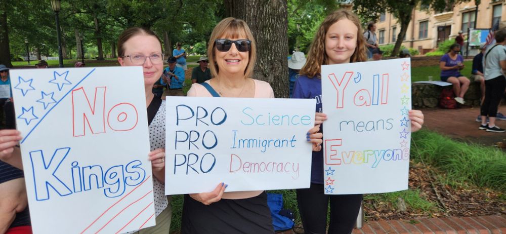 3 people holding signs at the No Kings protest in Chapel Hill. The signs say "No Kings" "Pro Science Pro Immigrant Pro Democracy" "Y'all means everyone"