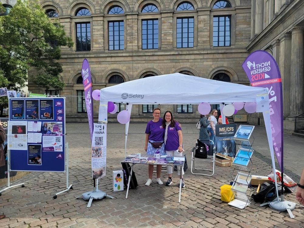 Zu sehen ist ein Infostand-Pavillon von der Patienteninitiative NichtGenesenKids.
Links befindet sich eine Schautafel, 2 Fahnen stehen am Pavillon, ein Ständer mit Infobroschüren, einige Plakate, ein kleiner Campingtisch mit weiteren Infomaterialien. Ein paar Luftballon in passenden NichtGenesen Farben (lila-Töne) hängen am Pavillon. 2 Frauen mit lila NichtGenesen Shirt stehen unterm Pavillon und beantworten gerne Fragen.