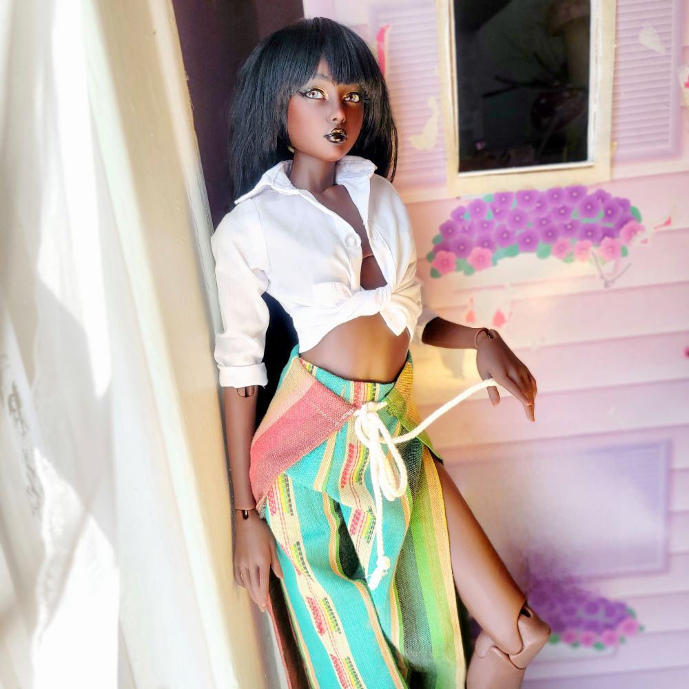 Dark skin Dollshe Diana with dark hair in a white front tie shirt and a colorful pair of wrap pants. 