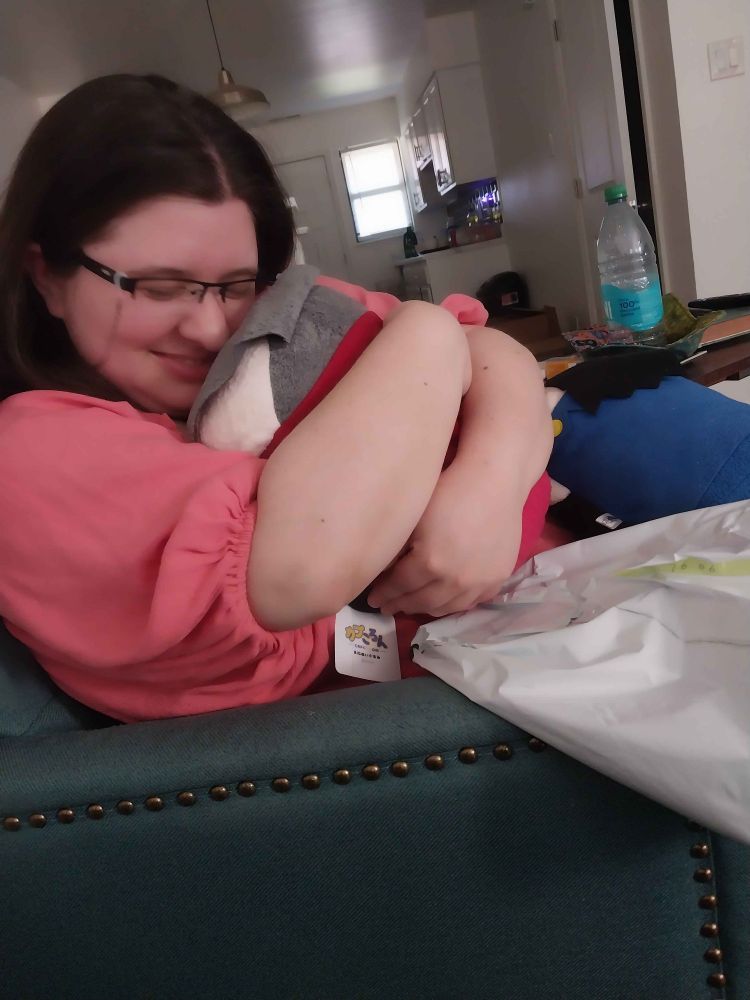 OP, a trans man wearing a salmon pink dress, is smiling very fondly and cradling a medium-sized Miles Edgeworth tsum tsum in his arms. Medium Phoenix Wright tsum tsum is also visible off to the side.