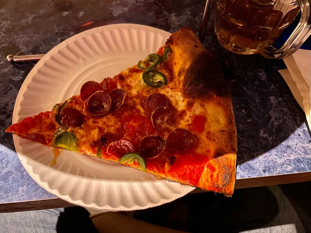 A slice of the pizza on a paper plate. Beer off to the side 