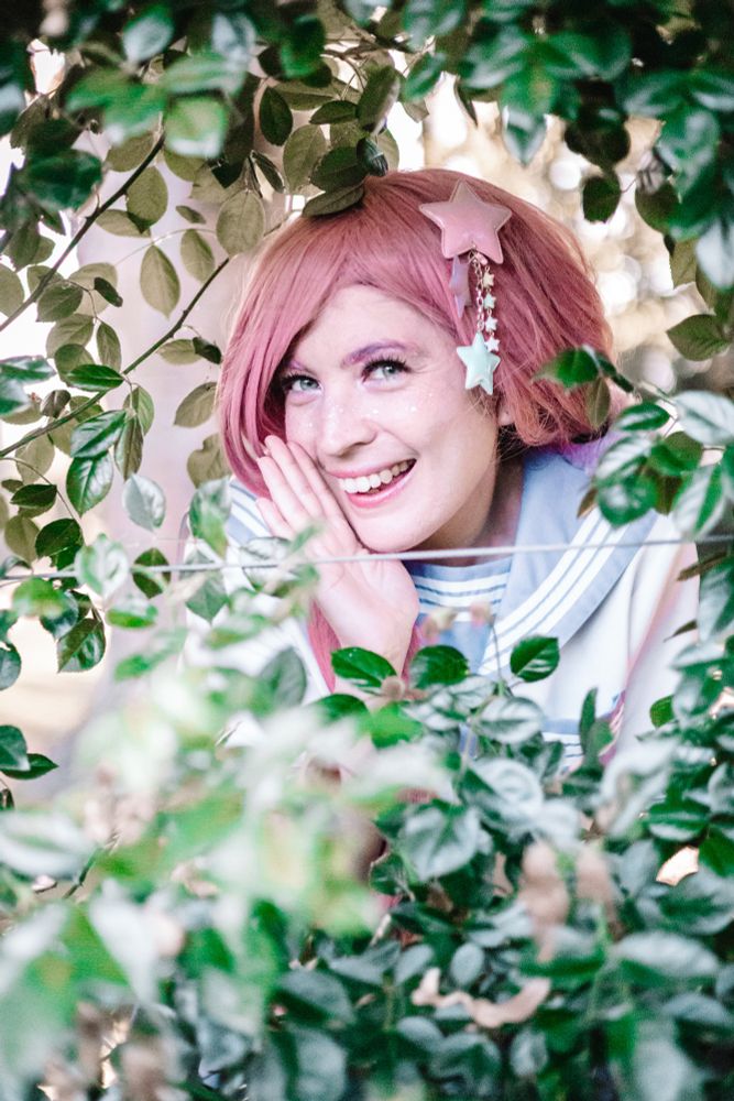 A photo of me cosplaying Luka Megurine in her After School model from the project diva games. I'm holding my hand up to my face like I'm going to share a secret, and I'm surrounded by leaves