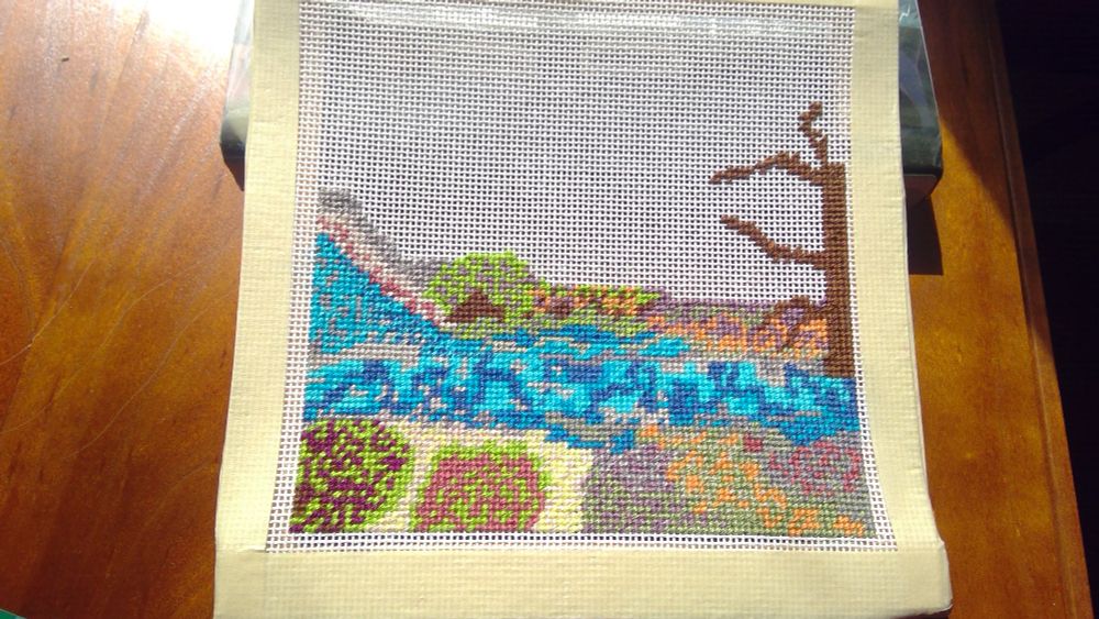Eight inch square needlepoint canvas about halfway filled. Scene has a river, flowers and greenery and rocks, and a dead tree.