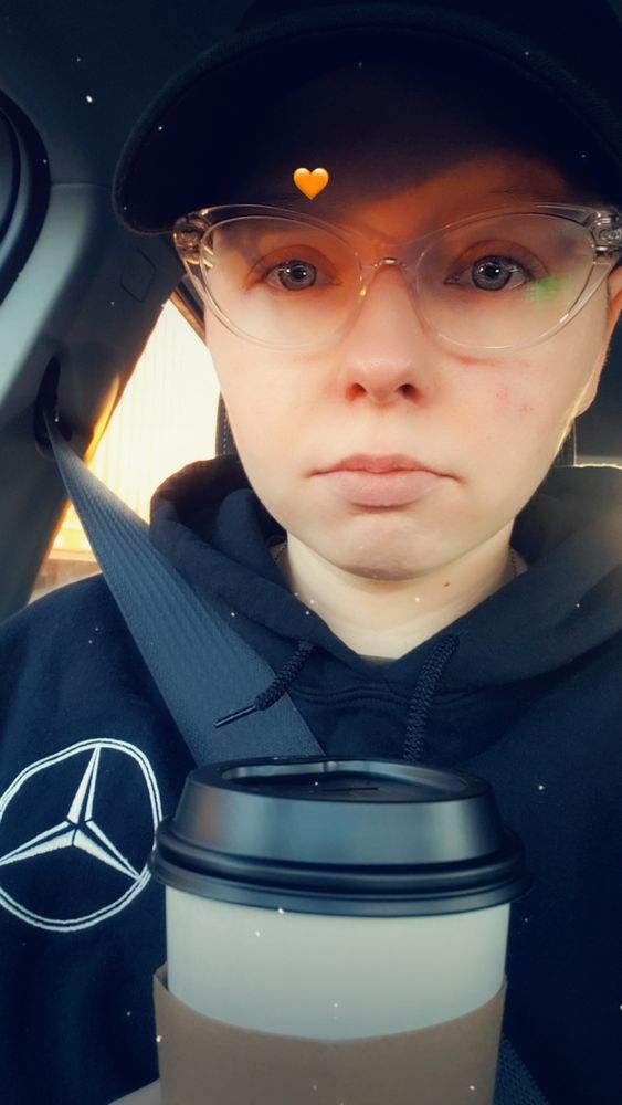 June looking tired in their car, holding a to go coffee. They're wearing glasses, a black ball cap, and a black Mercedes hoodie