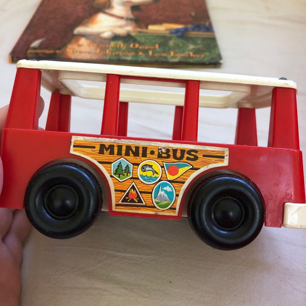 Vintage fisher price mini bus. Red plastic bus with black wheels and open white lid to add little people. Woodgrain sticker on side shows camping scenes (tent, pine trees, campfire, fish).