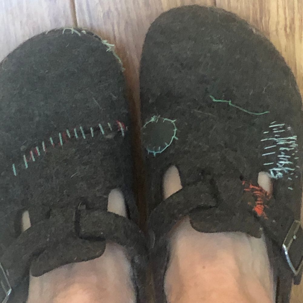 A repaired pair of charcoal grey felt topped birkenstock clogs with my (Caucasian & quite pasty this time of year!) feet in them. Left clog has turquoise stitching across the width in the middle of the toe section and also near the big toe. Right clog has a patched circle edged in turquoise stitch on the left side near the strap and messy turquoise stitching on the right with dark orange stitching on the strap itself on the right hand side.