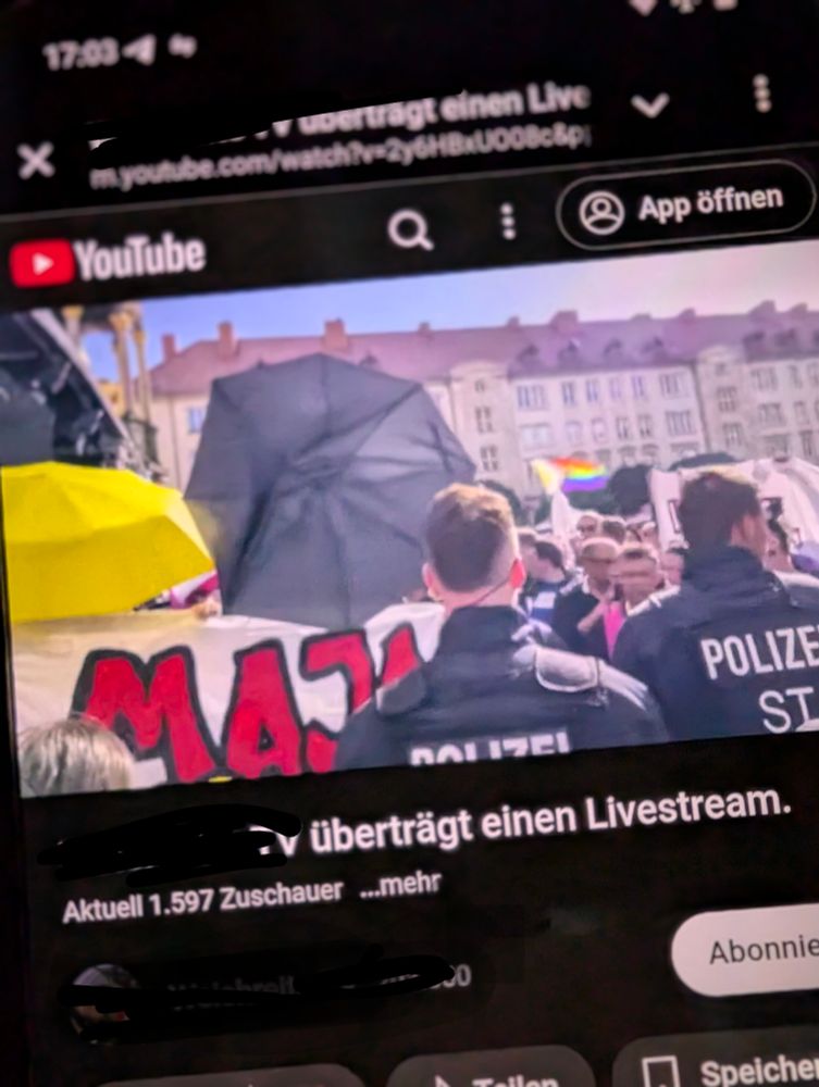 Ein Smartphone-Bildschirm zeigt einen laufenden YouTube-Livestream. Im Video sind Polizisten mit der Aufschrift „POLIZEI“ auf den Uniformen sowie mehrere Demonstrierende mit Bannern, Regenschirmen (schwarz und gelb) und einer Regenbogenfahne im Hintergrund zu sehen. Auf dem Banner ist in roten Buchstaben „MA…“ erkennbar. Oben im Display steht die Uhrzeit 17:03, unten wird angezeigt, dass aktuell 1.597 Zuschauer den Stream verfolgen.