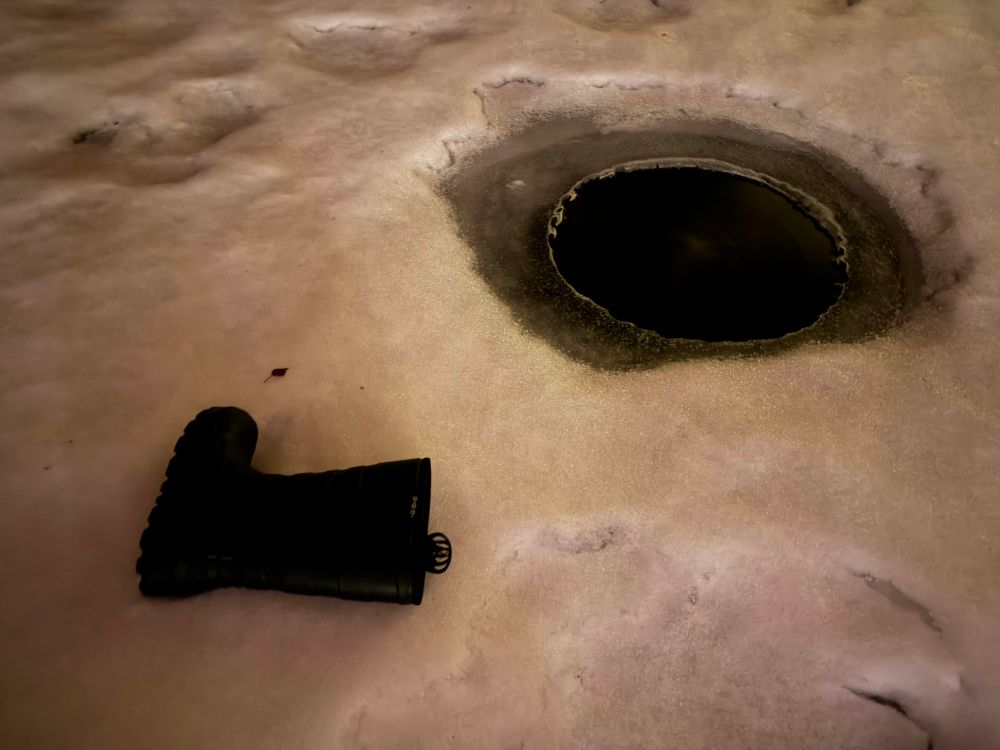 A rubber boot with a field recorder inside it next to a frozen pond with a hole in the ice.