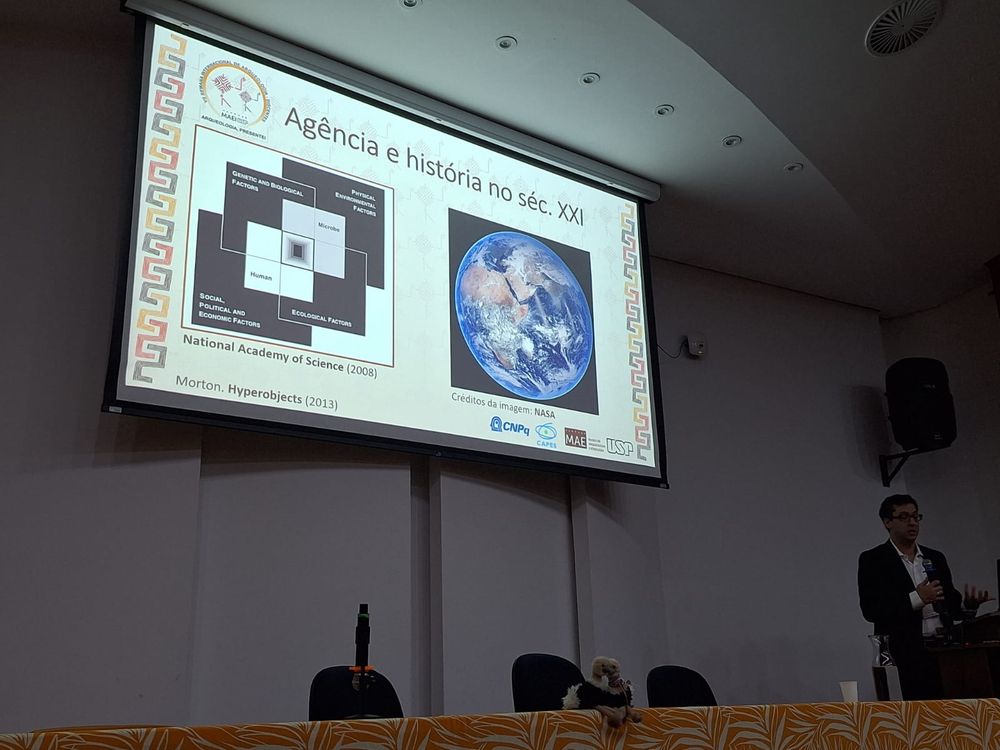 Vinicius Marino Carvalho discursando em um auditório. Nos slides, há uma foto do planeta Terra e um diagrama mostrando a intersecção entre fenômenos humanos e não-humanos no entendimento de epidemias. 