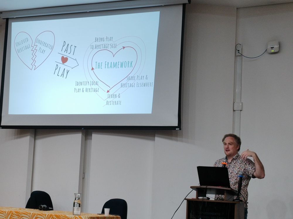 Professor Angus Mol discursa atrás de um púlpito. Seu slide mostra um coração partido, representando patrimônio e atos lúdicos malquistos, restaurados em um coração pleno pela iniciativa "Past, Love and Play" 