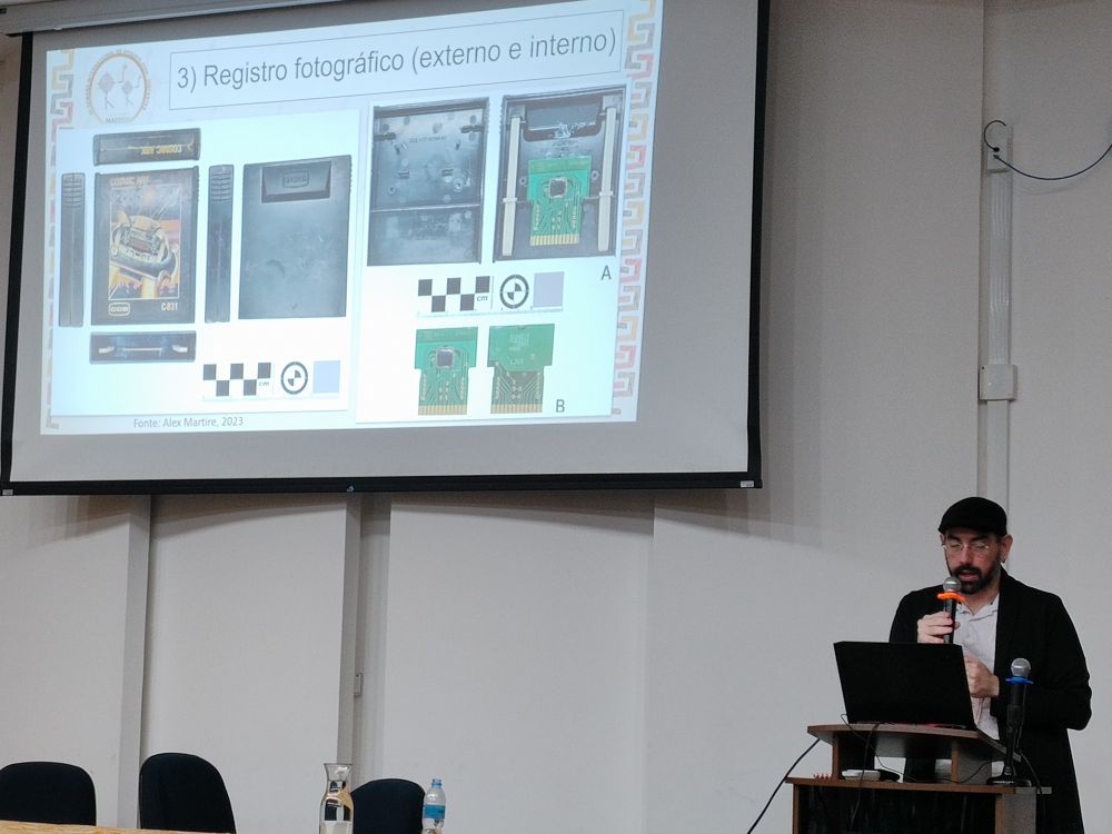 Professor Alex Martire discursa em auditório. Nós slides, vemos fotos de cartucho do Supergame 2800, tanto de sua parte externa quanto da interna. 