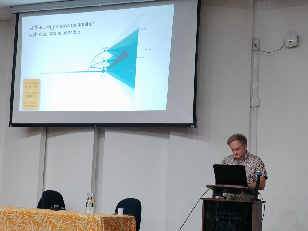 Professor Angus Mol fala atrás de um púlpito. Seu slide diz "Arqueologia mostra que outro caminho foi e é possível" 