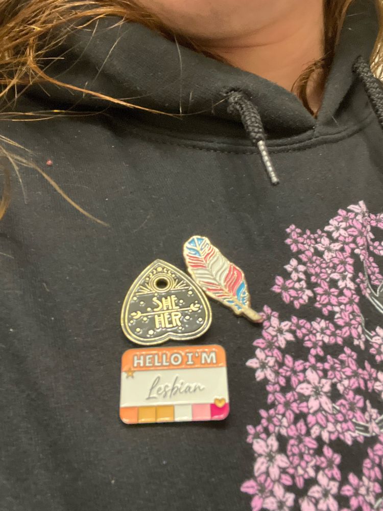 Close up on the feather, she/her, and lesbian pin. 