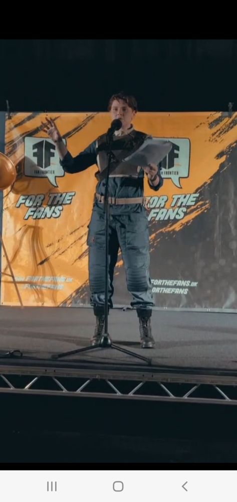 A person in a cal kestis cosplay from jedi fallen order standing on stage with a microphone in front of an orange back drop saying "for the fans"