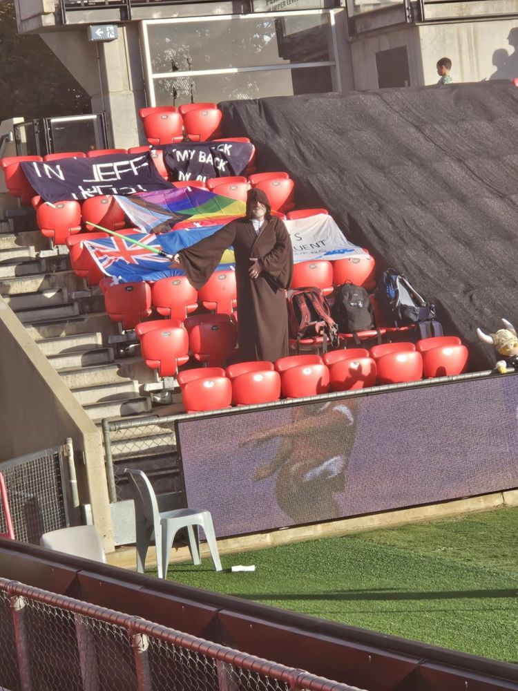 A man with a white beard in a brown Jedi cloak and holding out a green lightsaber standing in the away bay at Coopers Stadium. There are  7 rows of 6 seats exposed, to the right a black covering separates this bay from the rest of the stand. There are banners reading "In Jeff We Trust" and "My Neck, My Back, My Alex Chidiac", a progress Pride flag and two other obscured across the sear. It's half time. Everyone else is at the bar...