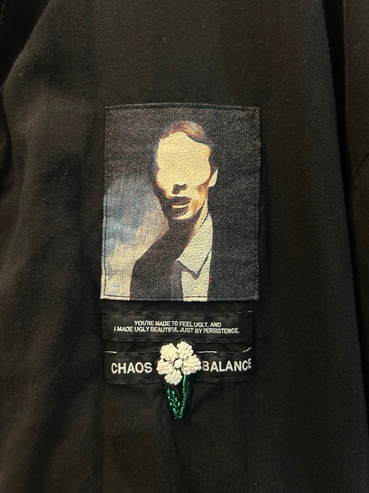 Undercover Jun Takahashi shirt detail: a painting of a white suited man with no eyes. A John Lyndon Quote embroidered beneath it: “You’re made to feel ugly, and I made ugly beautiful. Just by sheer persistence” and the words CHAOS BALANCE with a daisy. 
