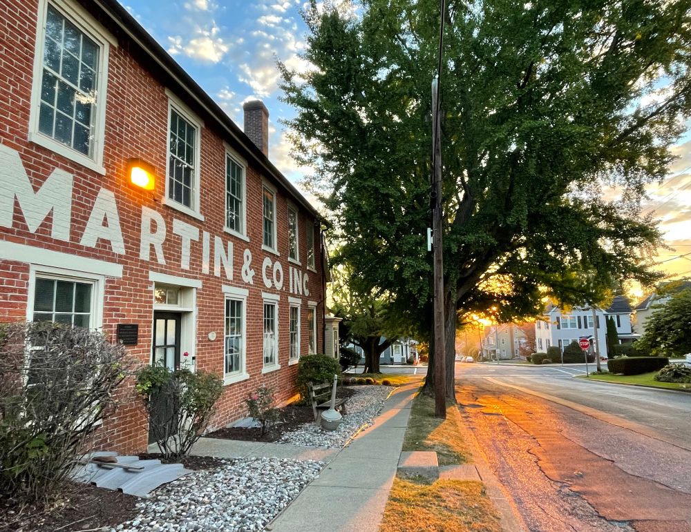 A wide angle shot showing an old brick building on the left with window frames a cream color and the name Martin & Co painted neatly between the upper floor and lower floor’s windows. Some shrubs and loose stones along the building are bordered by a sidewalk. Between the sidewalk and a macadam street a little further downhill are some large ginkgo trees still looking dark since the sun is just rising. Orange light from the rising sun seems to stain the road a glowing orange. There’s an intersection and beyond it nice small town houses and an orange-yellow glow low on the sky. What’s visible of the sky further up is blue with pretty small white clouds. Somewhere down near the golden glow is my home. 