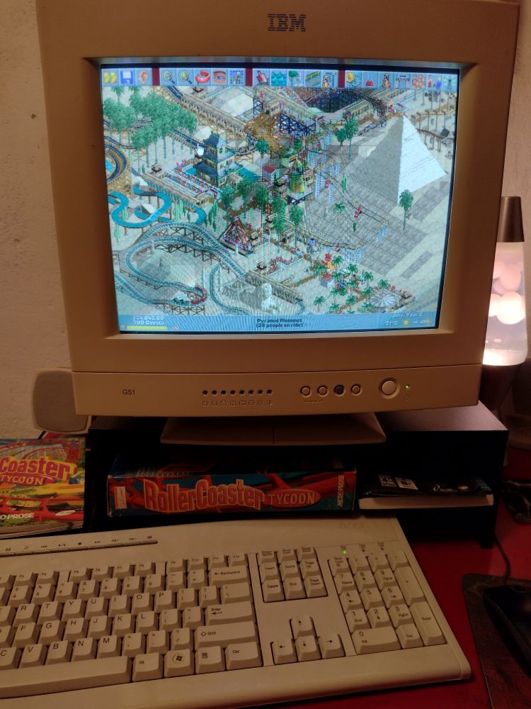 Rollercoaster Tycoon running on a CRT monitor. The game box and a lava lamp are visible to either side of the screen. The game is running the Dynamite Dunes scenario.