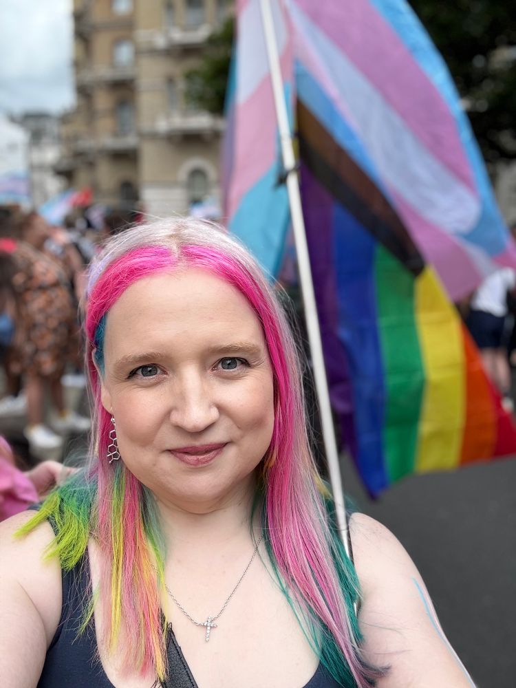 A woman with multicoloured hair with pride flags behind her, stood in the middle of the street.