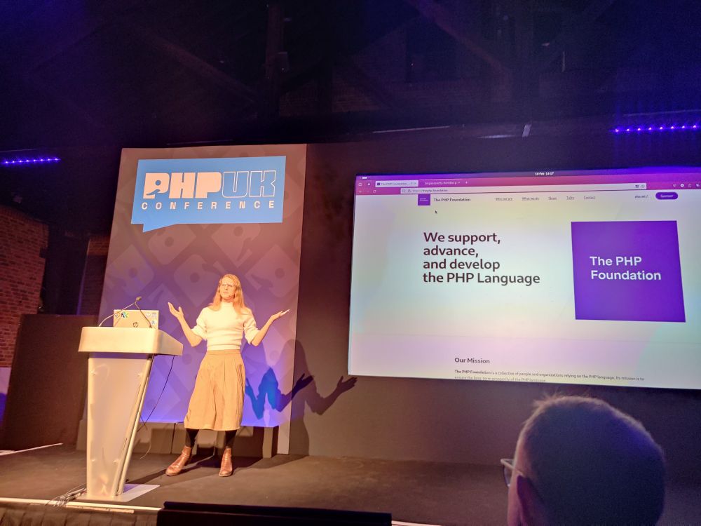Gina stood on stage in front of a screen showing the PHP Foundation website.