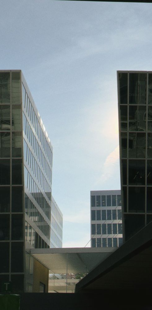 Two tall, glass-covered office buildings create a canyon-like space, framing a view of the blue sky with a few wispy clouds. In the background, a third building with a grid-like window pattern is visible. The architecture is sharp and modern, with reflections shimmering on the facades.