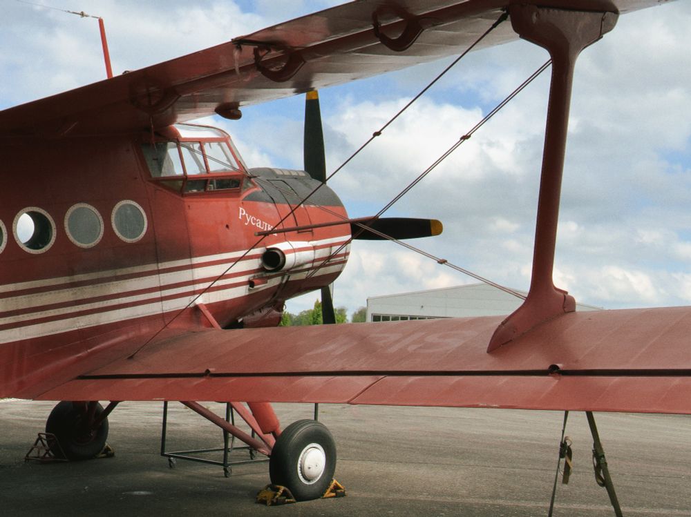 A red, vintage biplane with white stripes and Cyrillic lettering on the fuselage is parked on a tarmac. The aircraft’s double wings are supported by struts and wires, and the propeller stands still under a partly cloudy sky. A hangar building is visible in the background.
