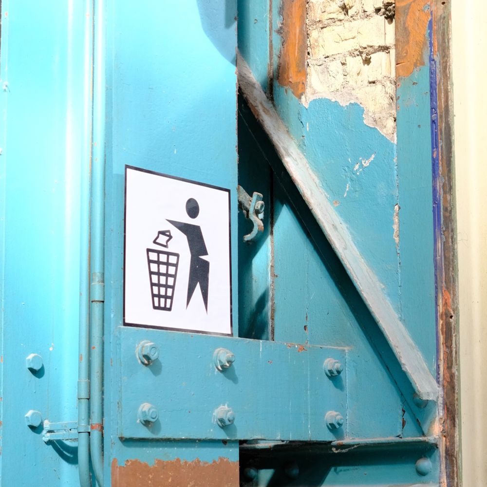 A turquoise wall, partially worn and stripped, reveals layers of paint and stone beneath. A simple black-and-white sign instructs people to dispose of trash properly, contrasting with the rough textures and vibrant colors of the background. The image has a gritty, urban character.