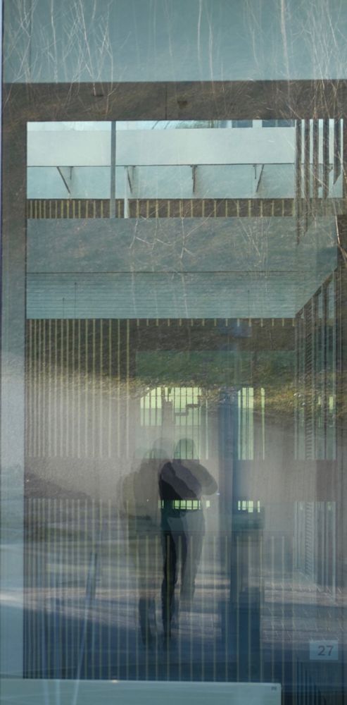 A photograph capturing an intricate interplay of reflections on glass. The overlapping layers of the building’s structure, trees, and the silhouette of a photographer create a fascinating optical illusion.