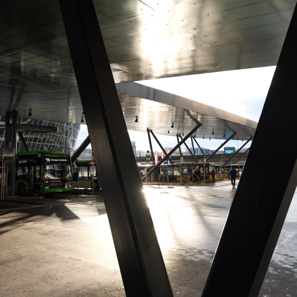 A sleek bus terminal featuring bold, V-shaped steel supports and a reflective metal ceiling. The morning or evening sun creates a warm glow and long shadows, casting a dramatic and cinematic mood over the mostly empty station.