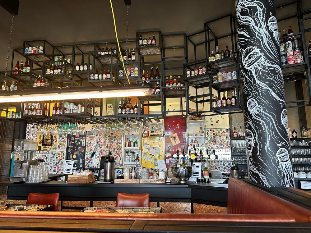 A stylish cafe&bar filled with shelves of liquor bottles, creative lighting, and sticker-covered walls. The eclectic decor, with a mix of modern and vintage elements, makes for an inviting and visually engaging atmosphere.
