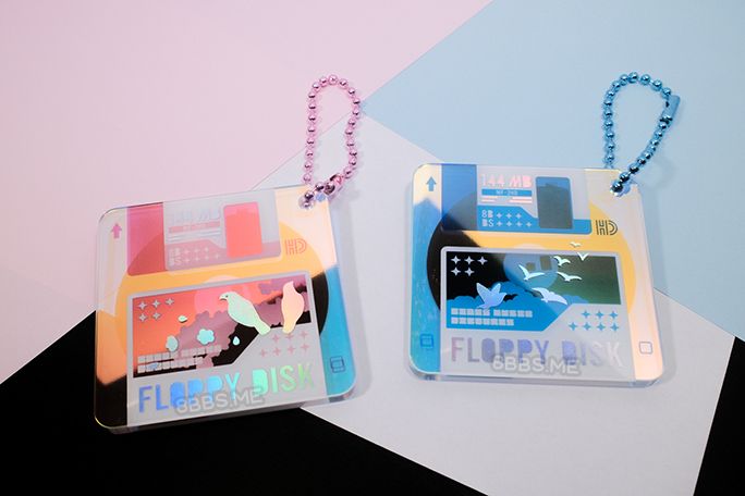 Photo of 2 acrylic charms styled to look like floppy disks. They are printed on rainbow acrylic with holographic silver foil accents and feature birds. The left one is pink with flowers, while the right one is blue and features clouds.