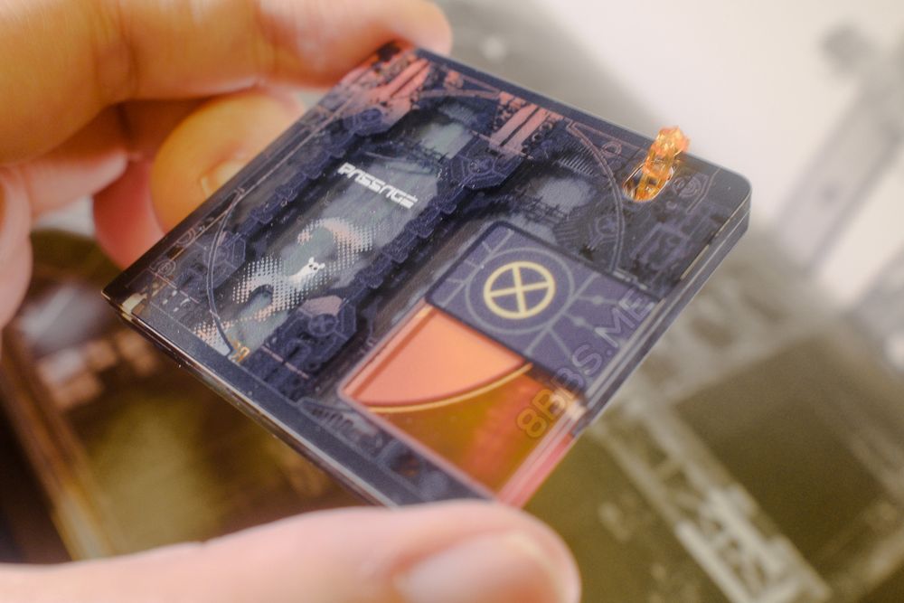 Closeup of an acrylic charm with layered printing for depth. The charm is shaped like a minidisc. It features a slugcat from the video game Rain World standing in an industrial gate, with the text "Passage" over its head. The charm is being held at an angle to showcase the depth of the layered printing.